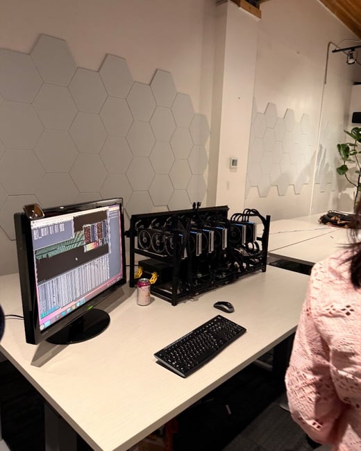 A photograph showing a cryptocurrency mining rig—an open frame housing multiple GPUs—sitting on a light-colored desk next to a monitor displaying technical logs. The background wall is decorated with white hexagonal panels, and the shoulder of a person in a pink sweater is visible on the right.