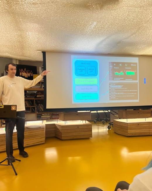 A man is giving a technical presentation indoors, standing next to a projection screen displaying a flowchart and performance graphs, while pointing at the diagram. The room features distinctive bright yellow flooring and wooden shelving in the background.