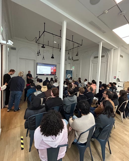 A wide-angle indoor photograph capturing a large audience seated in rows, attending a presentation or lecture in a spacious room with white walls and high ceilings.