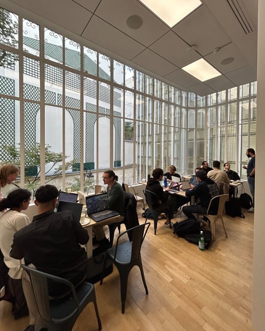 A candid photograph captures a group of approximately a dozen people working on laptops around tables in a bright, modern indoor space featuring a large glass wall overlooking a structured outdoor courtyard with green trellis patterns.
