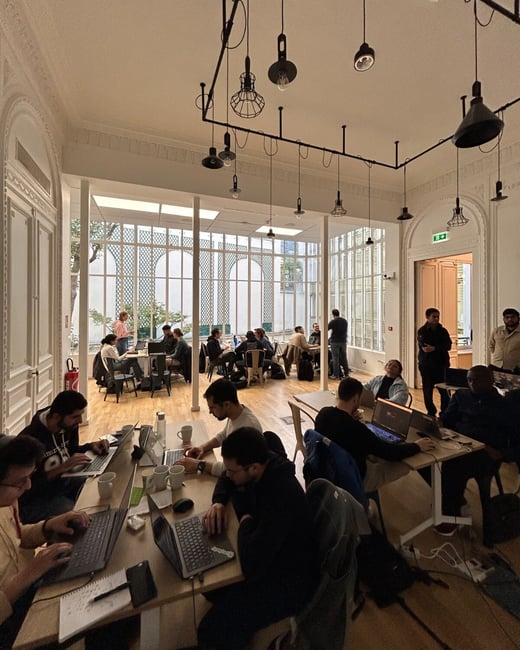 A candid photograph capturing a busy indoor work session, likely a hackathon or co-working event, set in a large room featuring ornate classical architecture and modern industrial lighting fixtures.
