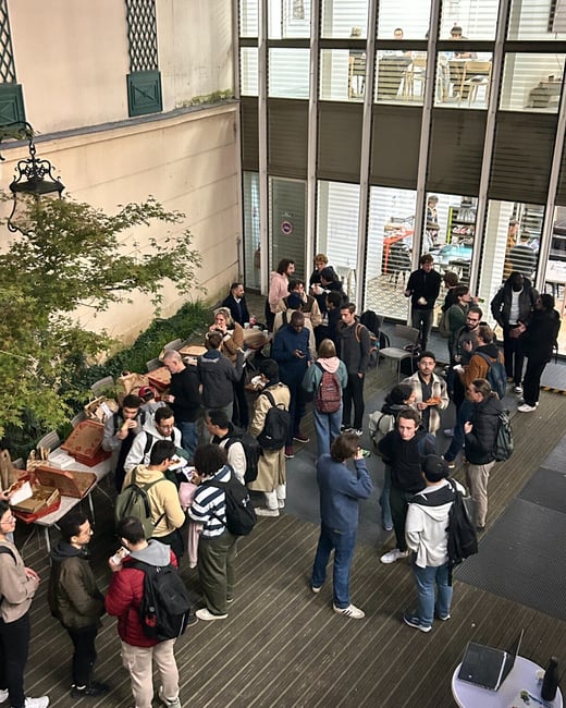 A high-angle photograph captures a large group of people gathered in an outdoor courtyard for a social event, eating food (likely pizza) next to a modern building with large glass windows illuminated from within.