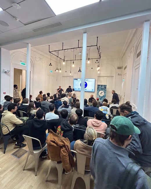 A photograph captures a large audience seated in a brightly lit, ornate white room, attending a presentation or panel discussion focused on a large screen at the front.