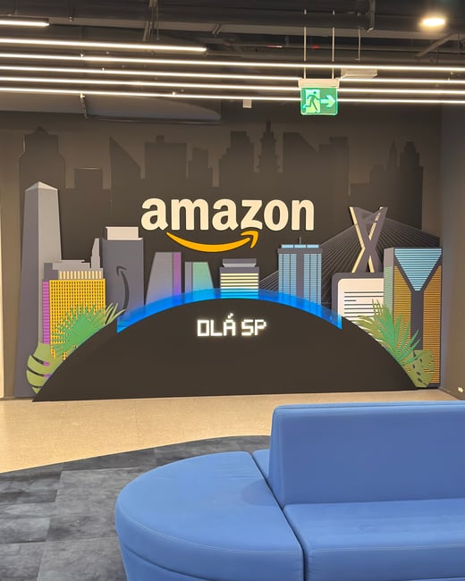 A photograph of an office lobby or reception area featuring a decorative wall with the Amazon logo, a stylized cityscape backdrop, and the text 'OLÁ SP' above a blue curved sofa.