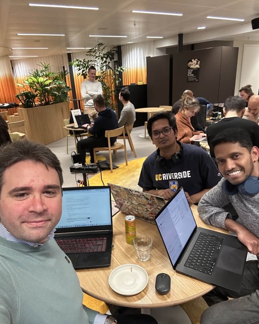 A group selfie taken in a modern, brightly lit office or co-working space, showing three men seated around a table with open laptops in the foreground, while numerous other people work in the background.