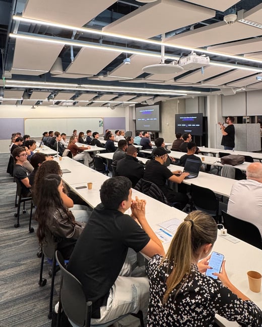 A vertical photograph capturing a presentation or lecture inside a modern classroom, with an audience seated at long white desks facing a presenter at the front.