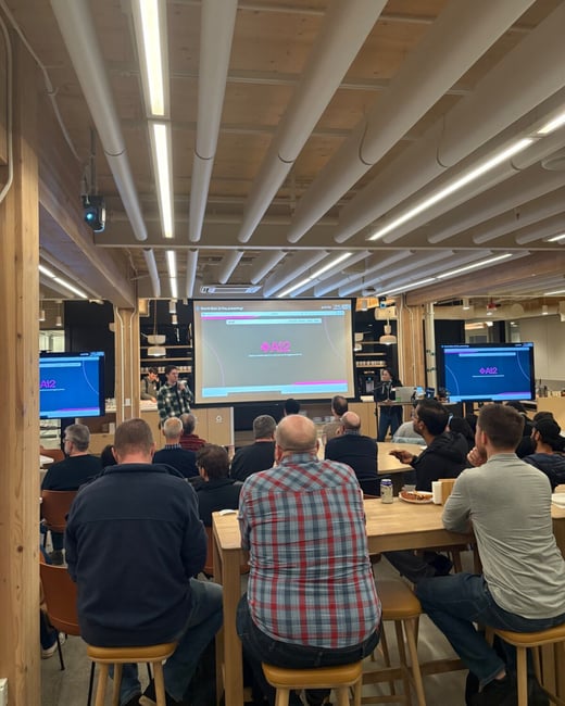 A candid photograph captures an indoor presentation setting where an audience is seated on stools and tables, facing a large projection screen and two side monitors. A presenter stands near the screen, which displays the AI2 logo and presentation title.