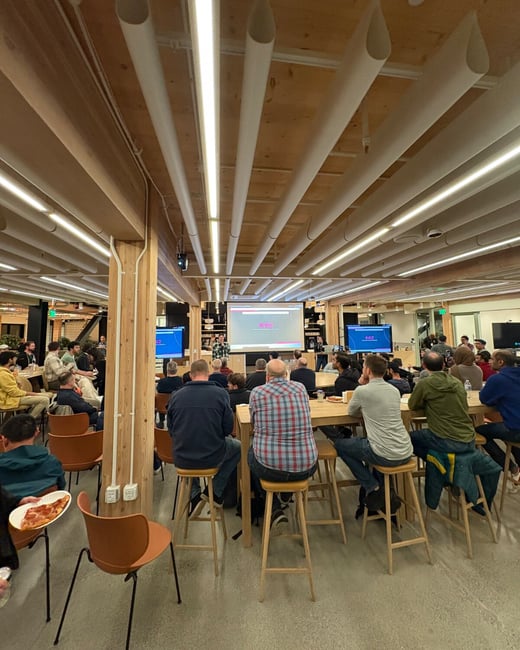 A photograph captures an indoor presentation or tech talk, showing a large audience seated on high stools and chairs facing a central projection screen displaying "SAIL" and two side monitors displaying "AI2," all situated beneath a distinctive wooden ceiling with long white light fixtures.