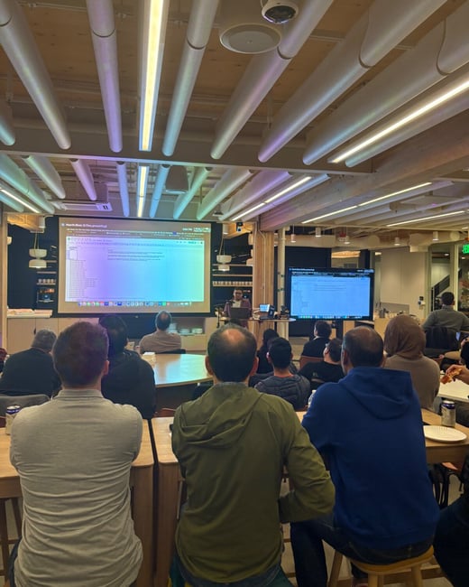 A photograph taken from the back of an audience showing a group of people attending an indoor presentation, where a speaker stands in front of two projection screens displaying data or code.