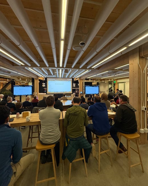 A photograph capturing a large group of people attending an indoor presentation or meeting in a modern venue characterized by high wooden ceilings and distinctive white cylindrical light fixtures, with attendees watching content displayed on a large central projection screen.