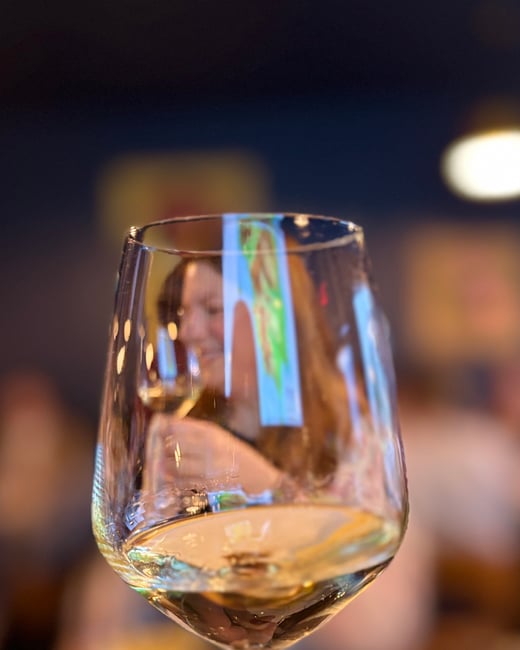 A close-up photograph of a hand holding a glass of white wine, with a blurred woman and a restaurant interior in the background.