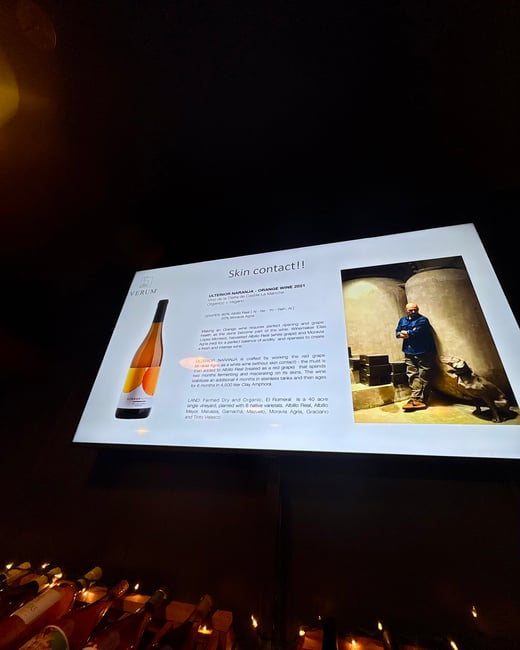 A photo of a presentation screen in a dimly lit room displaying information about 'Skin contact!!' orange wine, with wine bottles on a rack in the foreground.