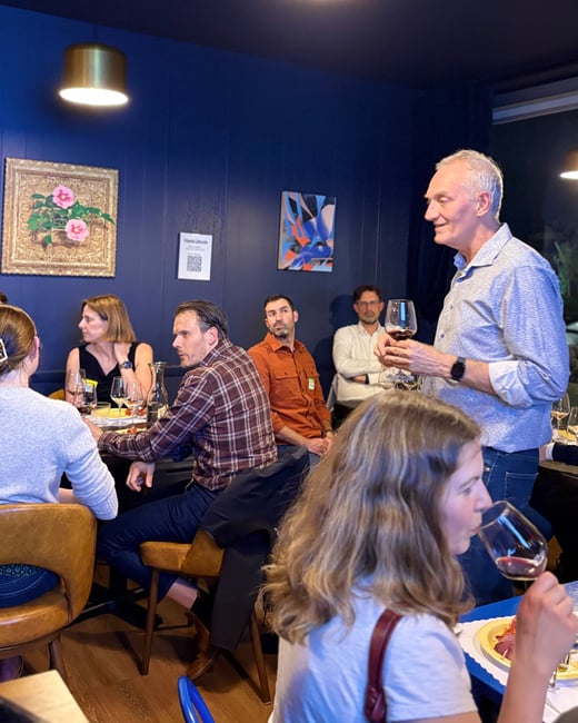 A group of people are gathered in a room with dark blue walls and gold pendant lights; one man is standing and holding a wine glass, possibly giving a toast.