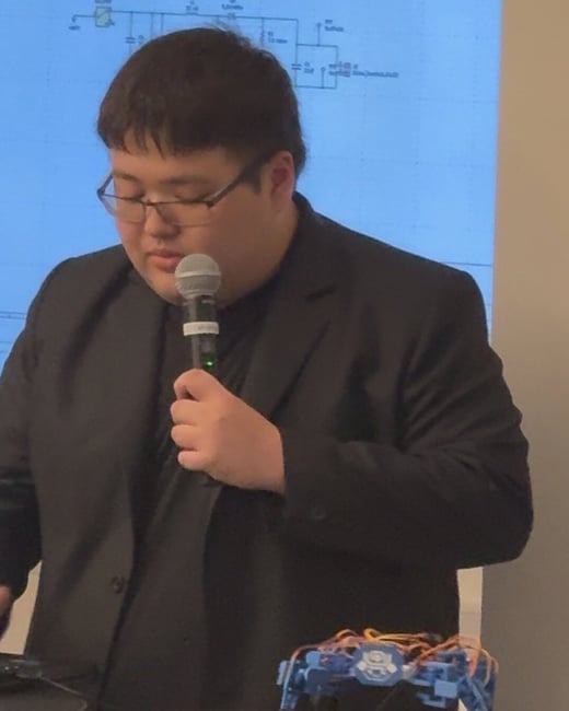 A man in a black suit jacket is giving a presentation indoors, speaking into a microphone while standing next to a table displaying a small, blue, multi-legged robotic device.