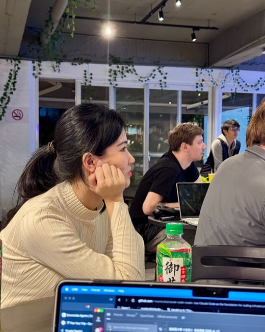 A candid photograph captures a group of people sitting at tables in an indoor space, likely a co-working area or cafe, with a woman in the foreground resting her chin on her hand and looking to the side. Several laptops and a green tea bottle are visible on the table.