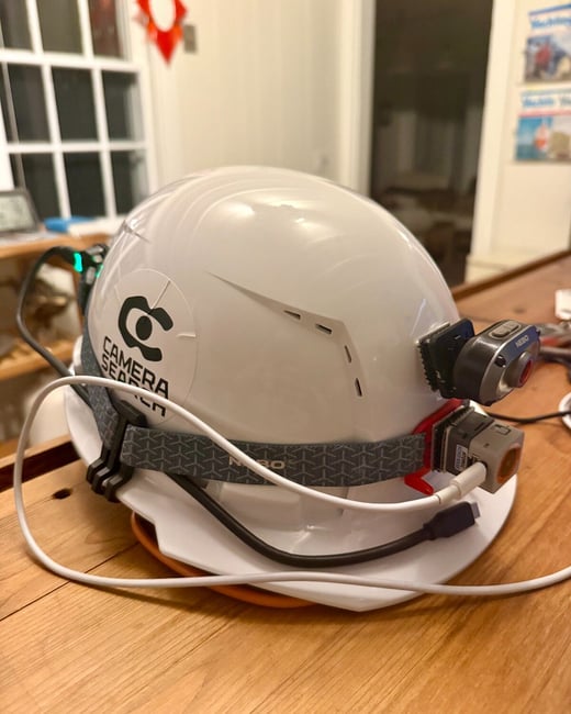 A close-up photograph shows a white hard hat resting on a wooden table, heavily customized with attached cameras, lights, and various black and white cables. The helmet features a black sticker displaying a stylized 'C' logo and the text 'CAMERA SEARCH'.