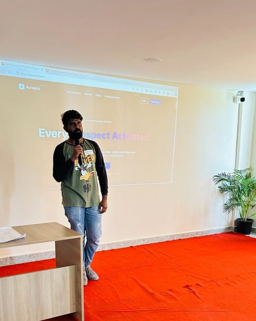 A bearded man is giving a presentation in an indoor venue, standing on a bright red carpet while holding a microphone. Behind him, a projection screen displays the homepage of the Kuralynx website.