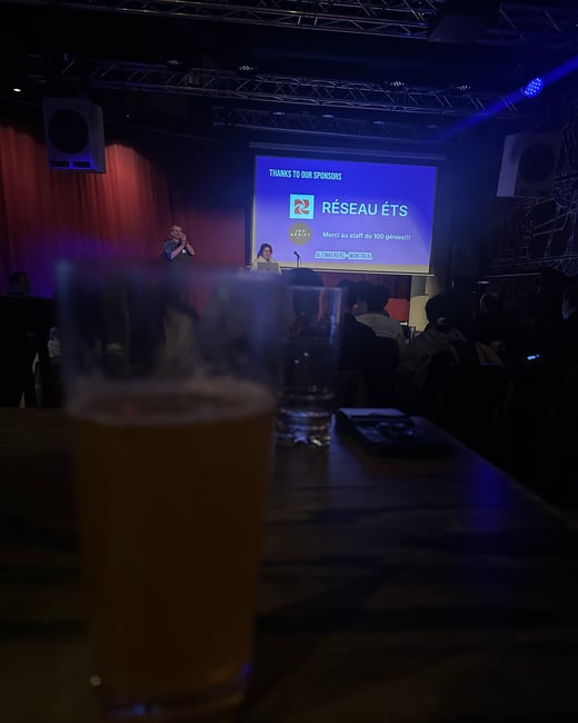 A dimly lit indoor event space featuring a presentation on a stage with a blurry glass of beer in the foreground.