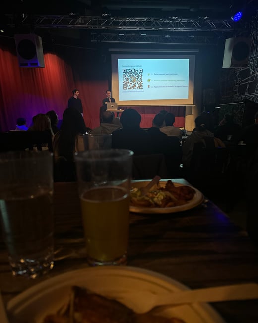 A photograph taken from an audience perspective of a technology presentation in a dimly lit venue, with food and drinks visible in the foreground.
