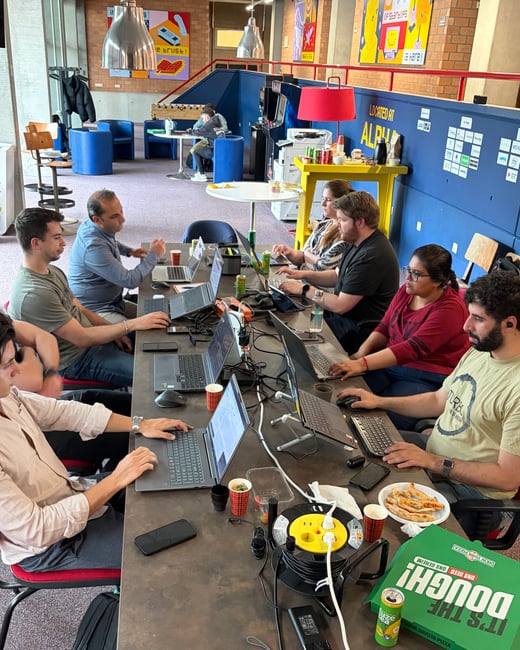 A group of people are gathered around a long table in a modern co-working space, focused on their laptops with pizza and drinks nearby.
