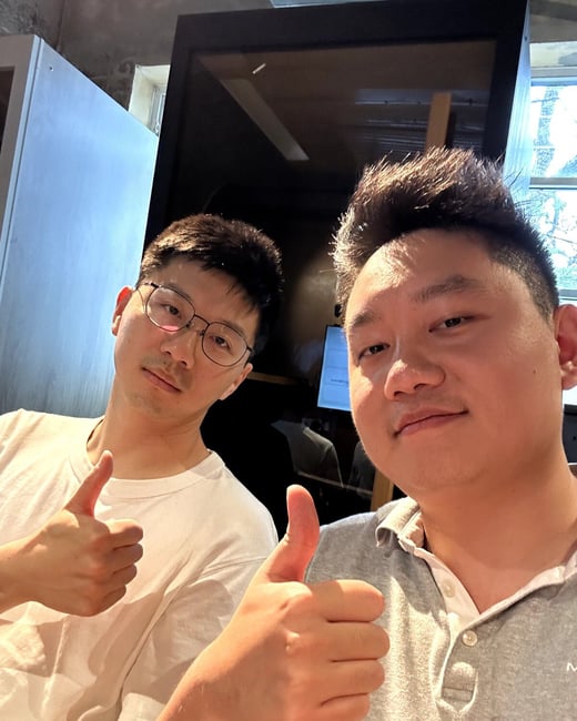 Two young men are posing closely together for a selfie indoors, both giving a thumbs-up gesture to the camera with expressions of approval or satisfaction.
