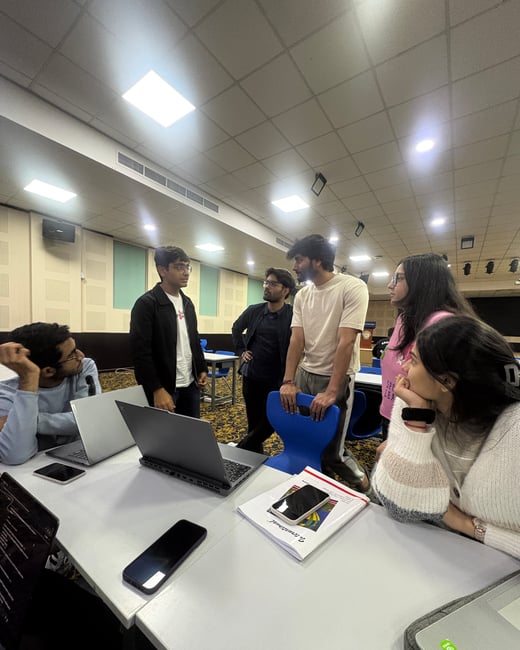 A group of young adults are gathered around a table in a large hall, engaged in a discussion with laptops and notebooks open.