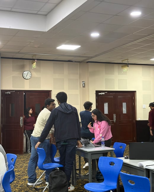 A group of people are gathered in a large room, likely a seminar hall, with some standing around a table with a laptop while others are near the entrance doors.