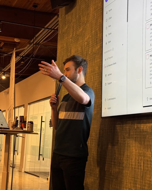 A candid indoor photograph shows a man giving a technical presentation, holding a microphone and gesturing toward a large display screen that shows a software application's user interface menu.