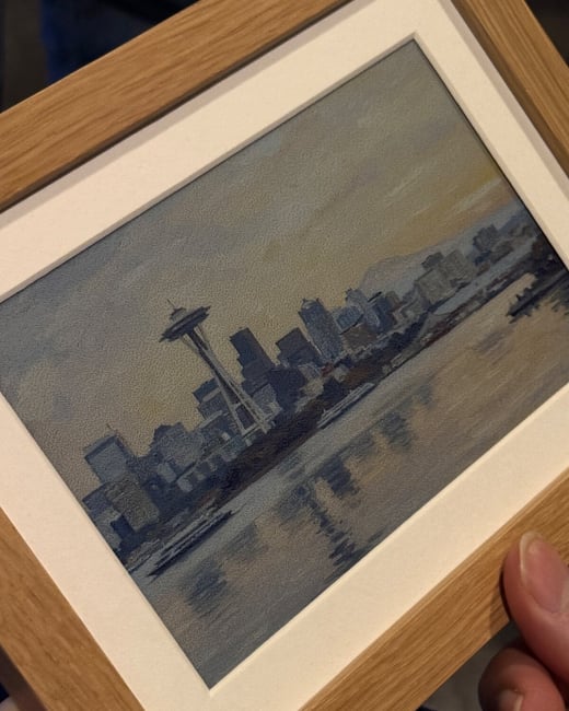 A close-up, angled photograph shows a person holding a small, framed artwork depicting the Seattle skyline, including the Space Needle, rendered in a muted blue and grey color palette with a textured or pointillist style.