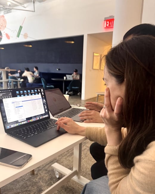 A woman in a tan sweater sits at a white desk in a modern coworking space, focused on her laptop screen while another person works beside her.
