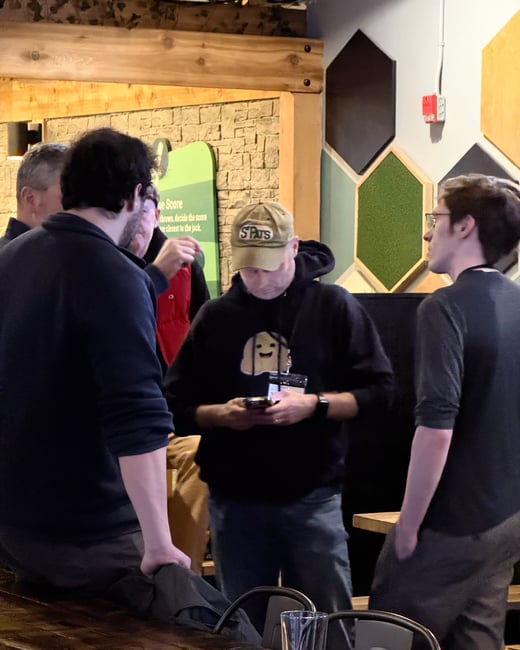 A group of men are standing and talking in a dimly lit indoor social space, with one man in the center looking down at his phone.