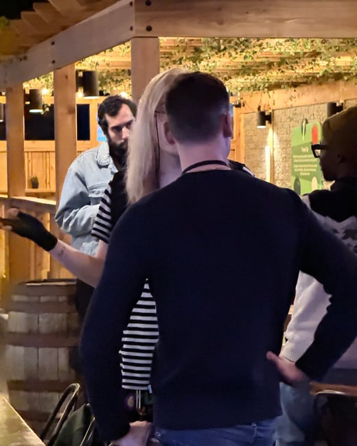 A candid photograph of a group of people socializing at an outdoor patio or bar area at night under a wooden pergola.