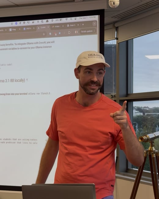 A man wearing an orange shirt and a white baseball cap is giving a technical presentation indoors, pointing toward a large projected screen displaying code and browser tabs.