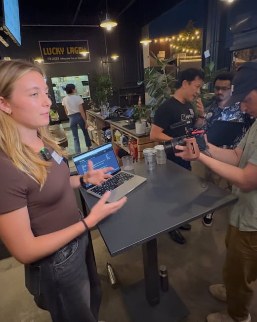 A candid photograph taken in a dimly lit indoor venue, showing a woman speaking and gesturing over a laptop while being filmed by a person holding a professional camera.