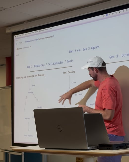 A man wearing a white cap and a red shirt is giving a technical presentation, pointing at a flowchart projected onto a large screen in an indoor setting. A Dell laptop sits open on a table in the foreground.