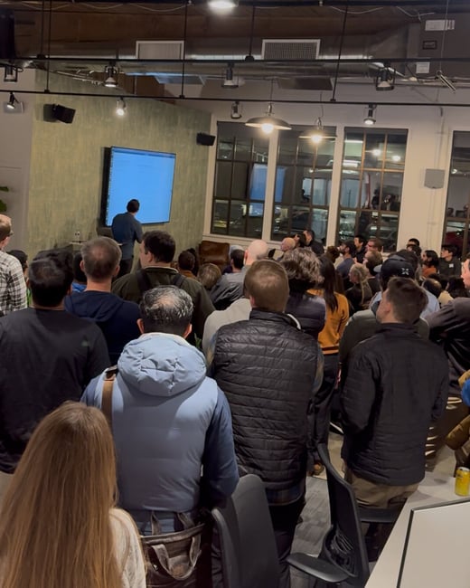 A photograph capturing a large audience gathered indoors in a dimly lit event space, watching a presenter standing in front of a large, brightly lit screen displaying blue content.