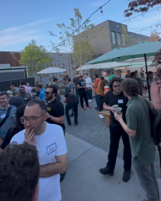 A candid photograph capturing a large group of people socializing and networking outdoors during an evening event in an urban setting, featuring string lights and industrial architecture in the background.