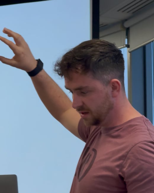 A man wearing a maroon t-shirt is giving a presentation indoors, gesturing with his right arm raised while looking down at a laptop in the foreground.