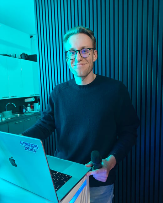 A man wearing glasses and a dark sweater stands behind a podium with a laptop, holding a microphone. The scene is illuminated with vibrant cyan and blue lighting against a backdrop of a slatted wall and a kitchen area.