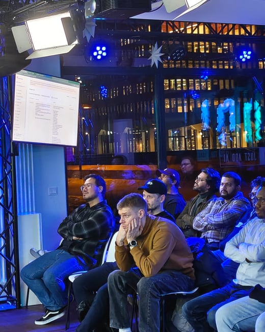 A group of people are seated in a dimly lit room, attentively watching a presentation displayed on a large screen.