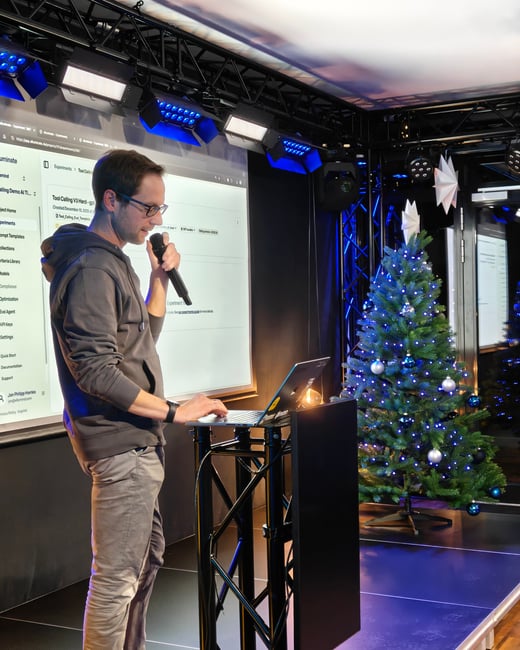 A man stands on a stage giving a presentation, holding a microphone and using a laptop. A large screen behind him displays a software interface, and a decorated Christmas tree stands to his right.