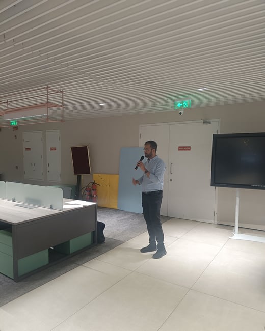 A man stands in a modern office or event space, holding a microphone as if giving a presentation or speech.