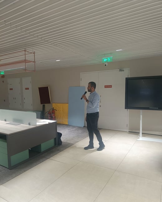 A man is standing in a modern office environment, holding a microphone as if giving a presentation or speech.