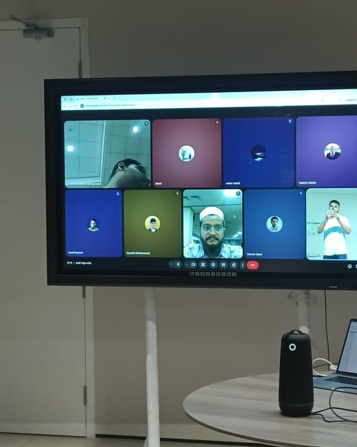 A photograph showing a large monitor displaying a Google Meet video conference with several participants, set in an office environment.