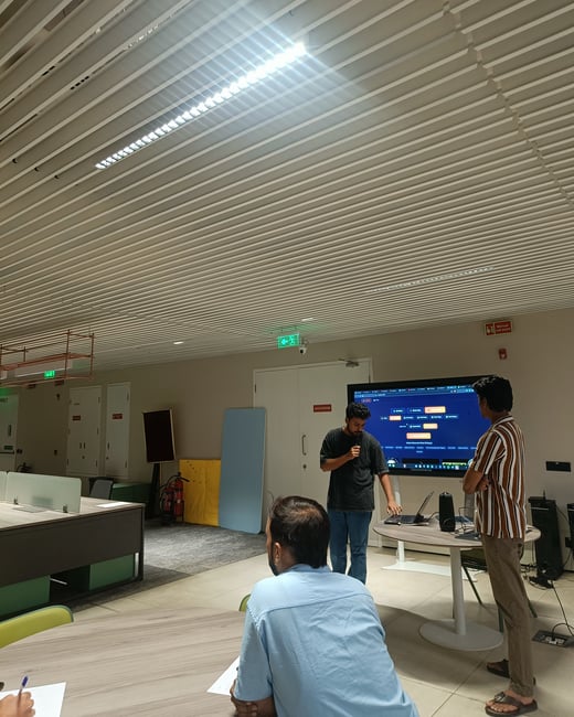 A man is giving a presentation or software demonstration in a modern office setting, using a large digital screen while others observe.