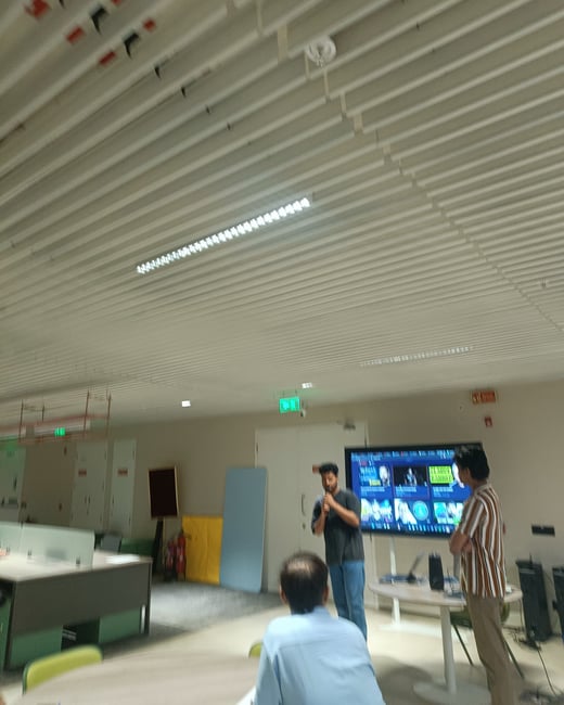 A man is giving a presentation in a modern office setting, standing next to a large display screen while a few others observe.