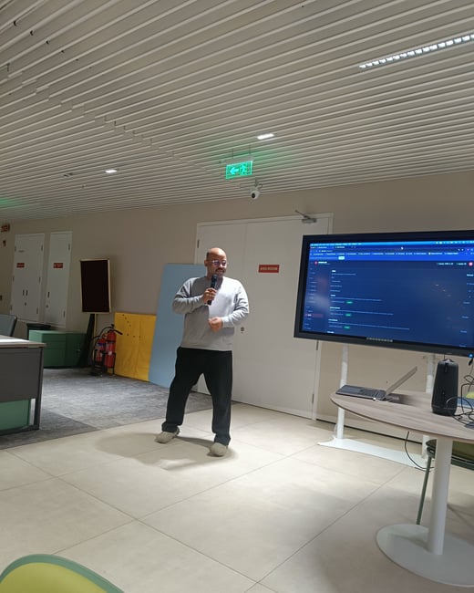 A man stands in a modern indoor space, holding a microphone and giving a presentation next to a large digital screen displaying a software interface.