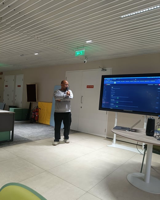 A man stands in a modern office or presentation space, holding a microphone and looking towards a large digital display showing a software interface.