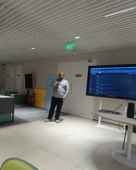 A man is giving a presentation in a modern office space, standing next to a large screen displaying a software interface while holding a microphone.