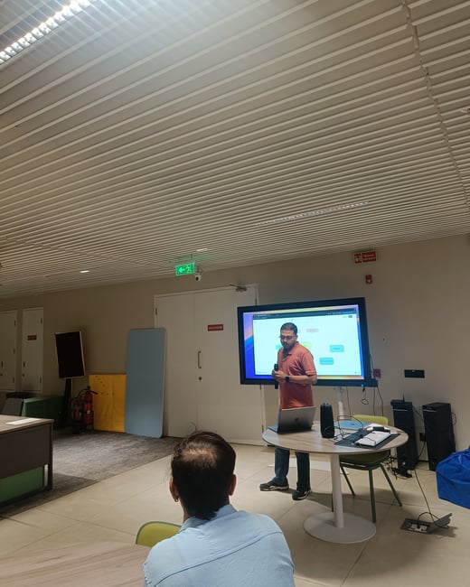A man is giving a presentation in a modern office setting, standing next to a large screen displaying a diagram and a table with a laptop.
